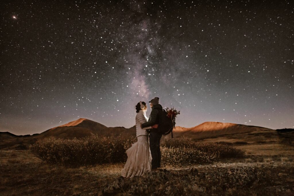 A man and a woman stand in a field with the starry night sky lit up above them.