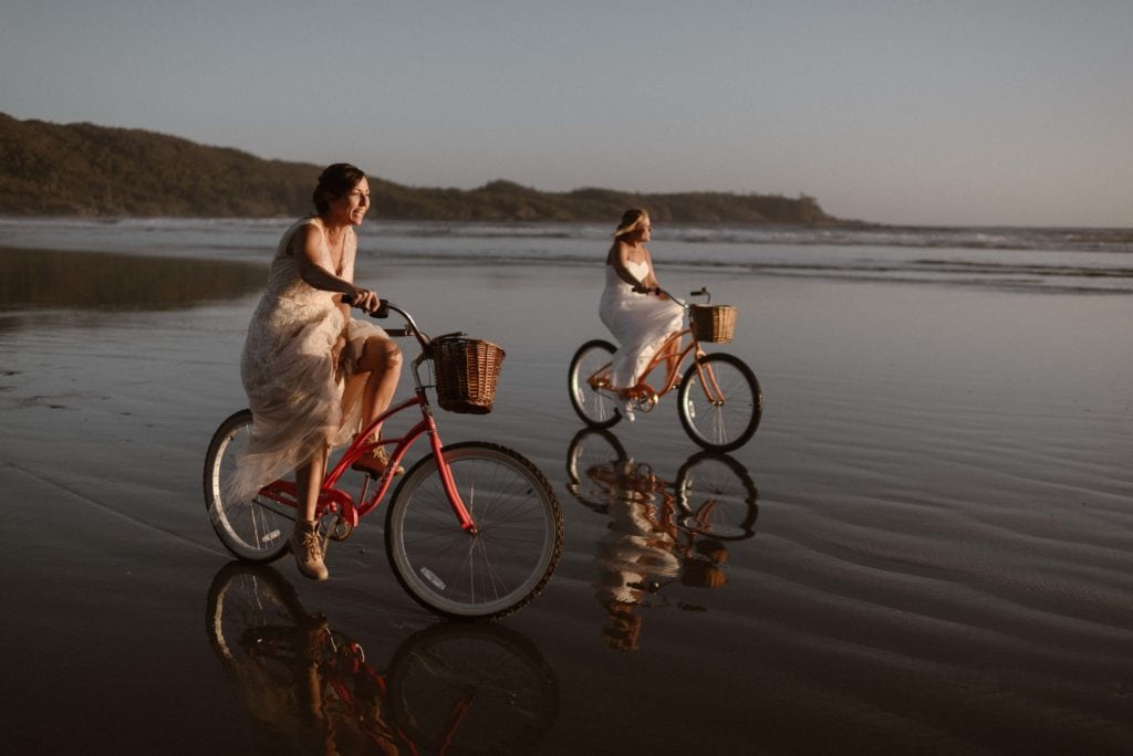 Two women in wedding dresses ride bikes on the coast.