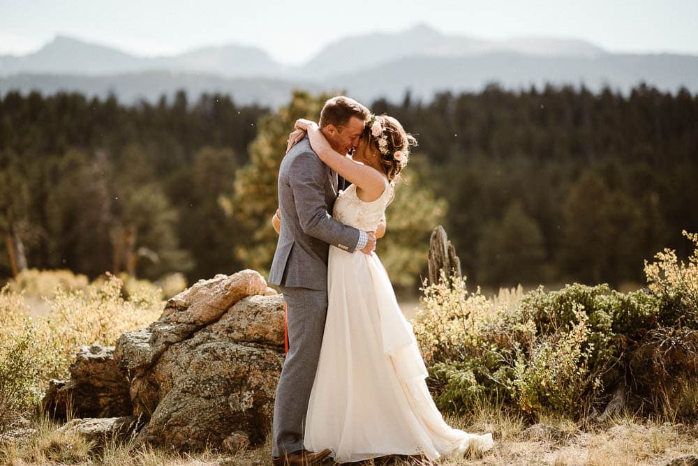 Bride and groom embrace in a meadow in Rocky Mountain National Park on their elopement day. There are forests and mountains behind them. 