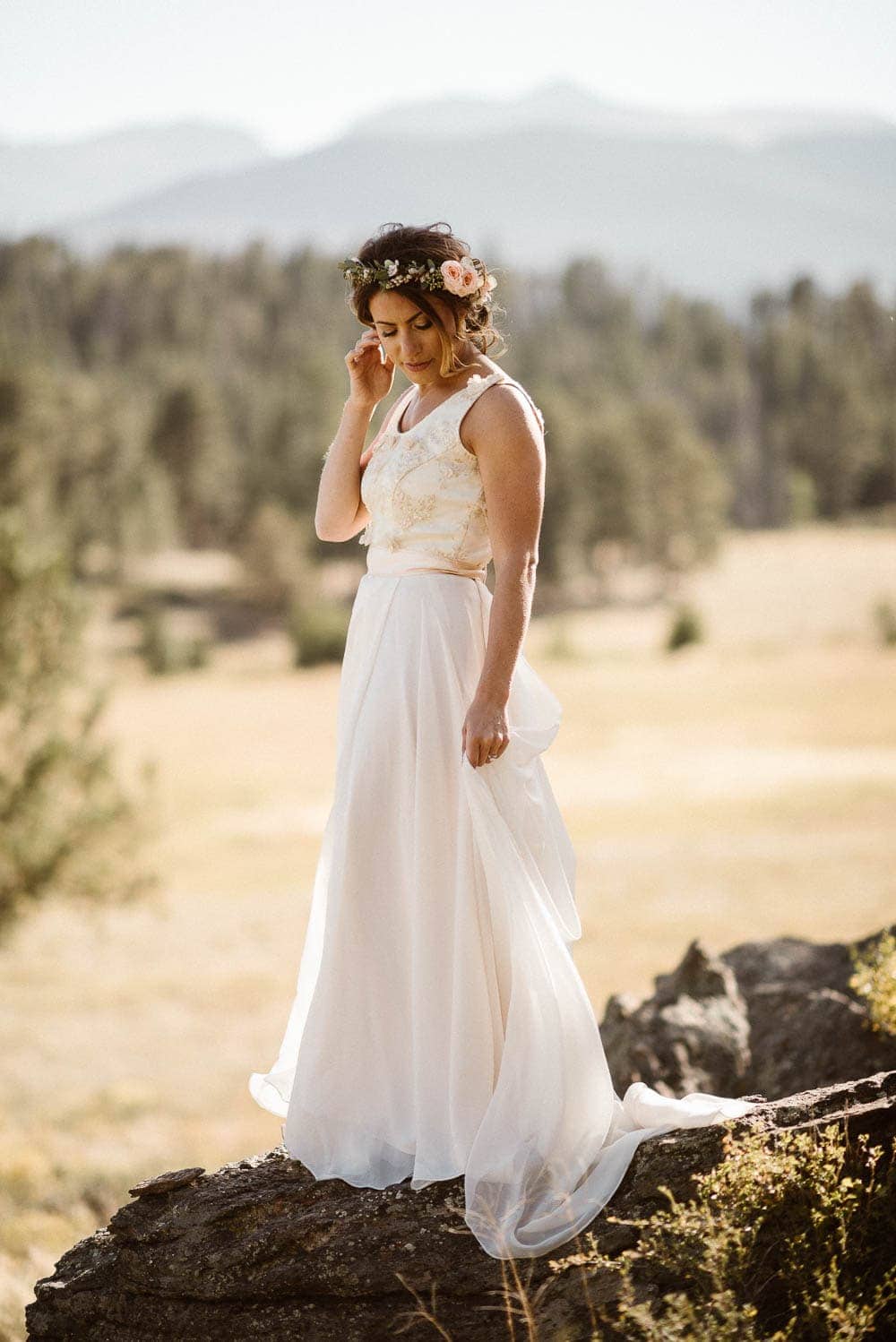 Bride stands on a rock and looks down, and holds the skirt of her white dress. She is wearing a flower crown with light pink roses.