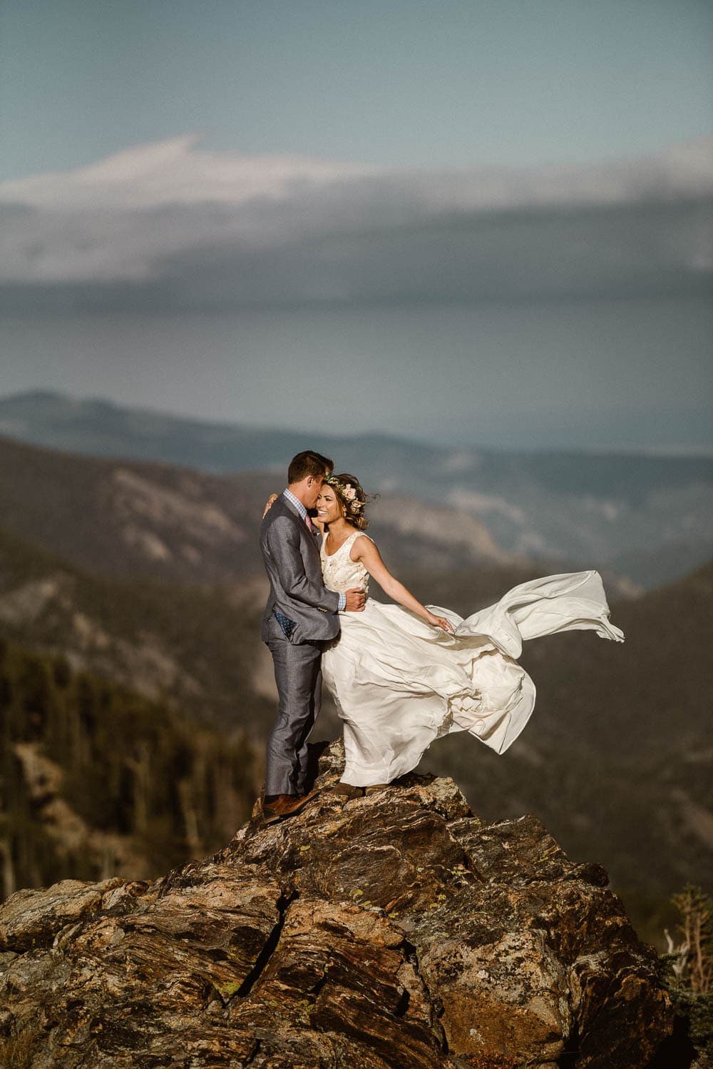 Bride and groom embrace, while bride's dress flows in the wind. There are mountains behind them in Rocky Mountain National Park. 