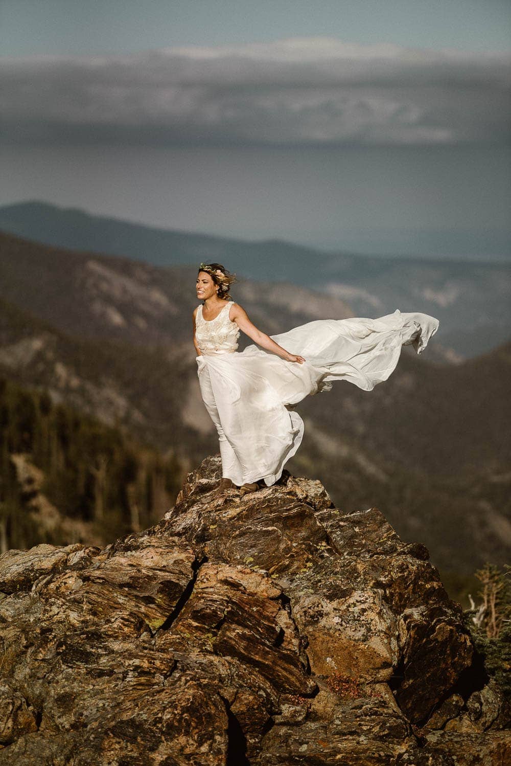 Bride stands on top of mountain, her white dress is flowing in the wind behind her. 