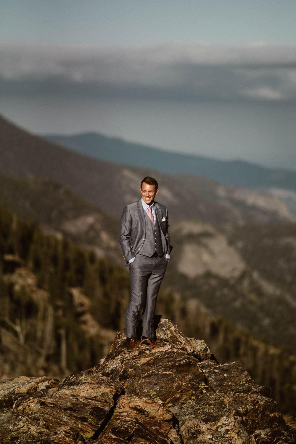 Groom stands on top of mountain, he is wearing a grey suit with a pink tie and smiling. 