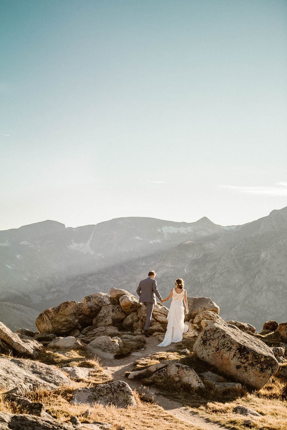 Bride and groom hold hands and climb up rocks, with mountains in the background, in Rocky Mountain National Park, Colorado. 