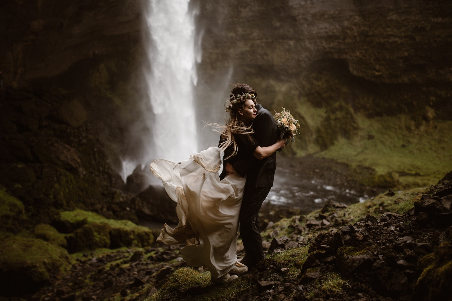 Bride and groom embracing in front of a waterfall in Iceland. The bride's white dress is flowing in the wind behind her. 