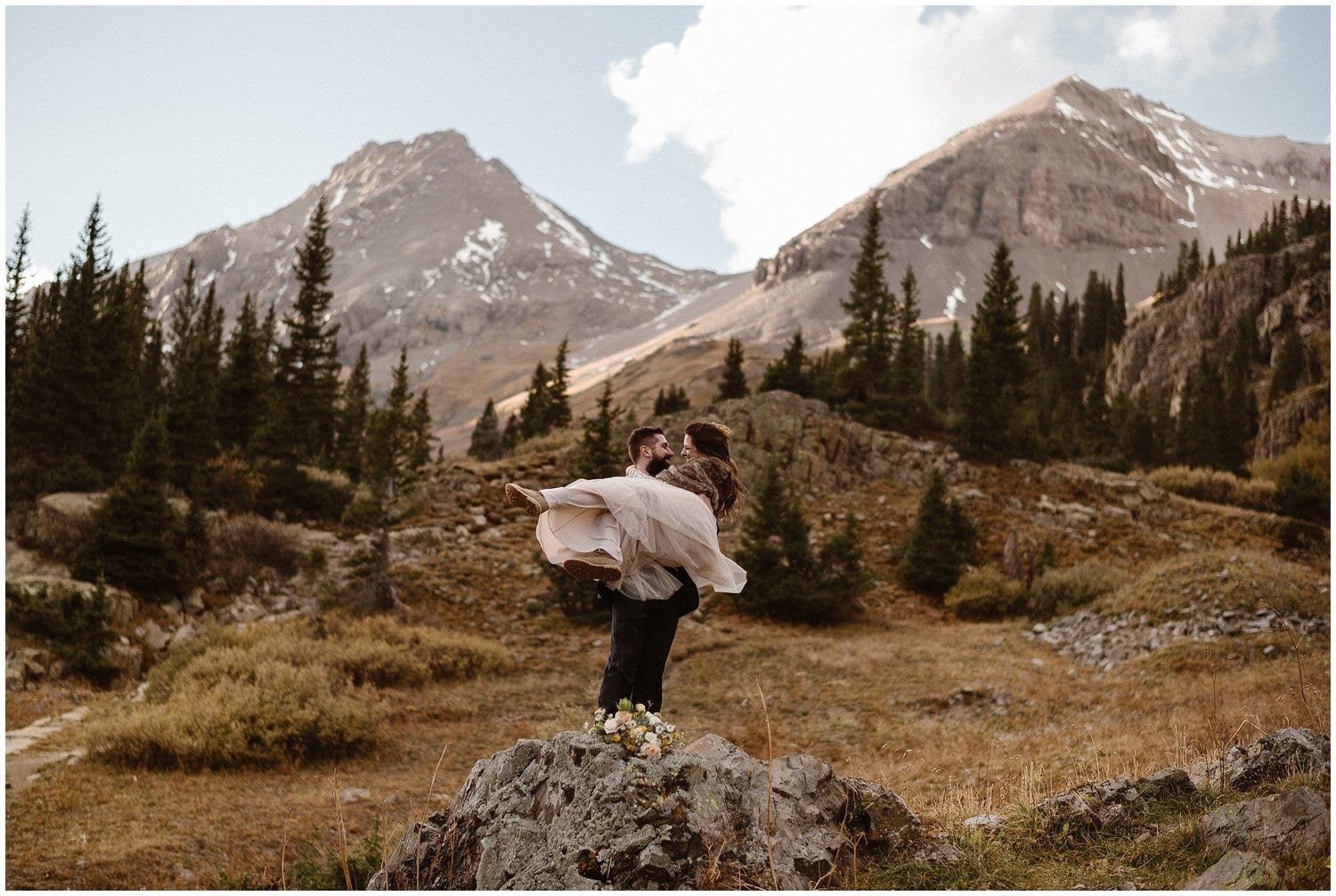 Groom lifts up bride in a meadow, with trees and mountains in the background. 