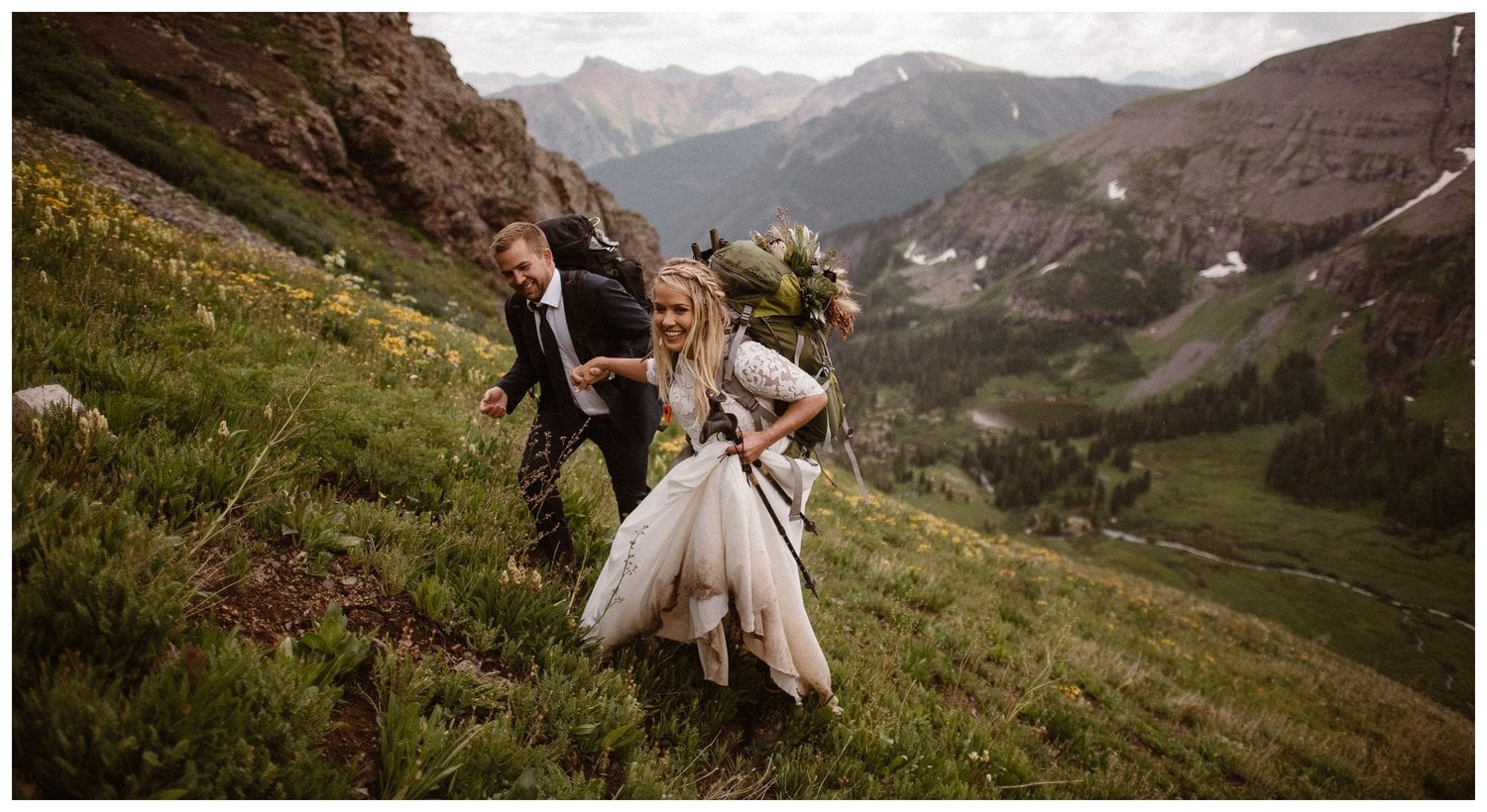 Bride and groom hold hands and climb up a grassy hill covered in yellow wildflowers. They are wearing wedding attire and there are mountains in the background. 