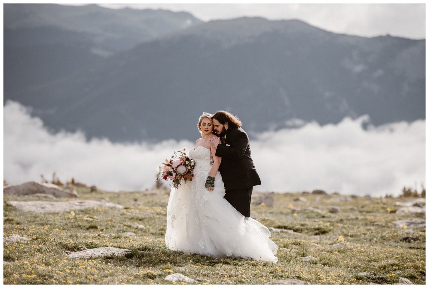 Bride and groom embrace in meadow with yellow wild flowers at Trail Ridge Road in Rocky Mountain National Park, Colorado. There are cloud inversions behind them. 