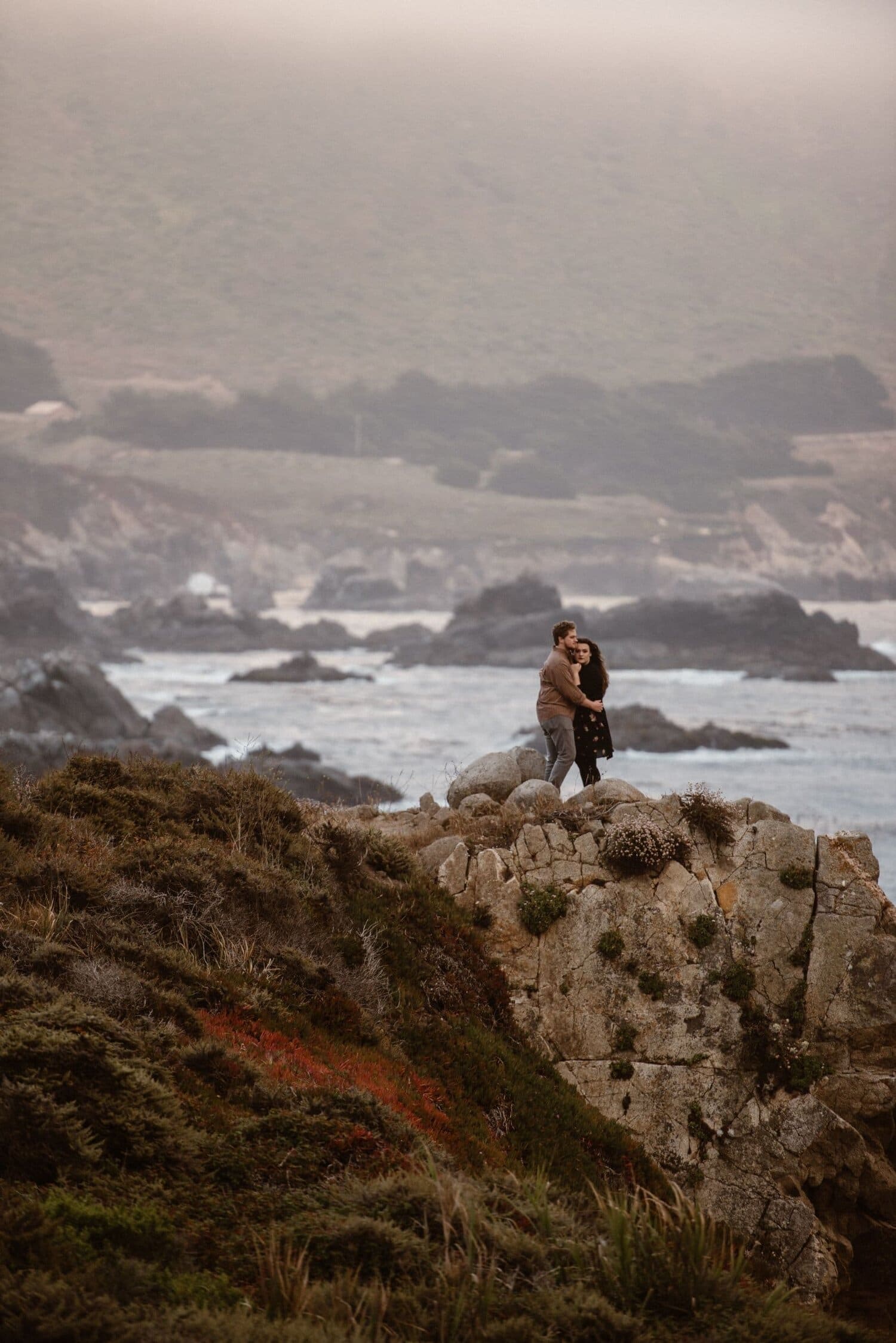 Bride and groom embrace at the top of a rocky cliff overlooking the ocean in Big Sur, California. 