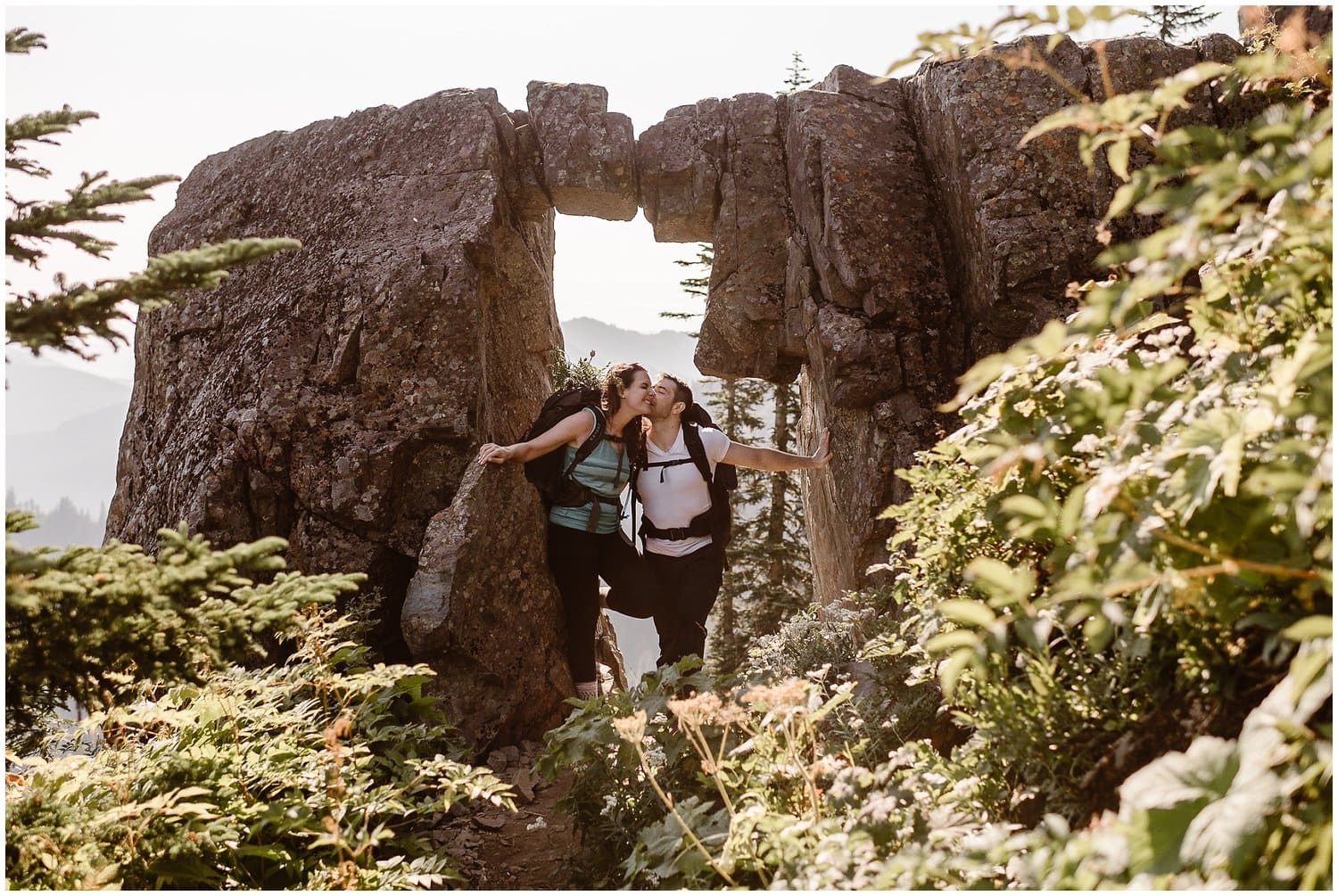 Bride and groom standing underneath a rock formation while hiking in Washington. The groom is kissing the bride's cheek.