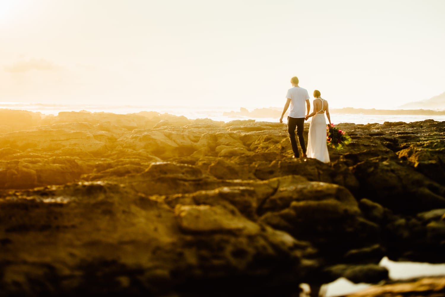 Bride and groom hold hands and walk across rocks along the shore in Santa Teresa, Costa Rica.
