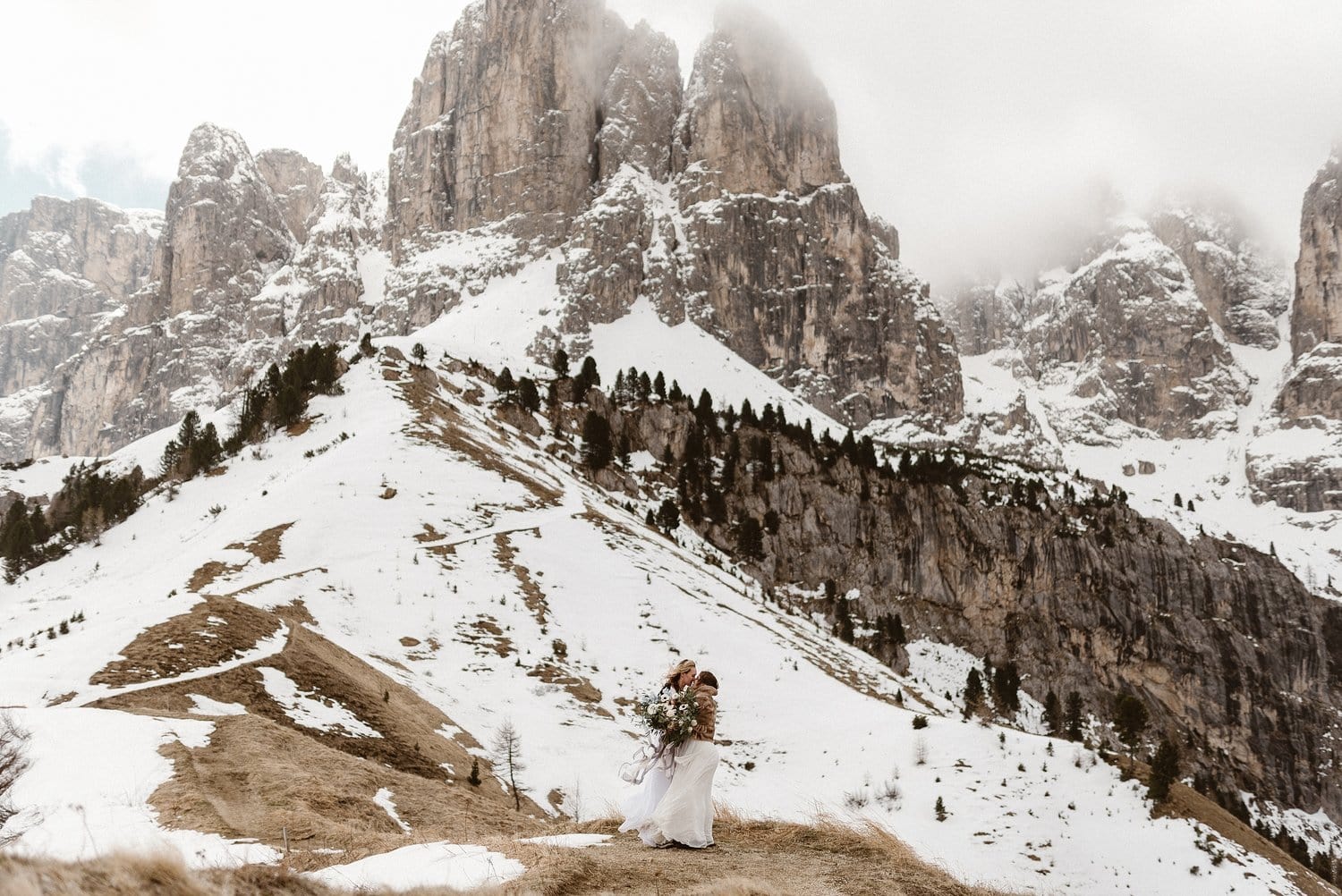 Two brides kiss in front of the snow-covered Italian Dolomites. 