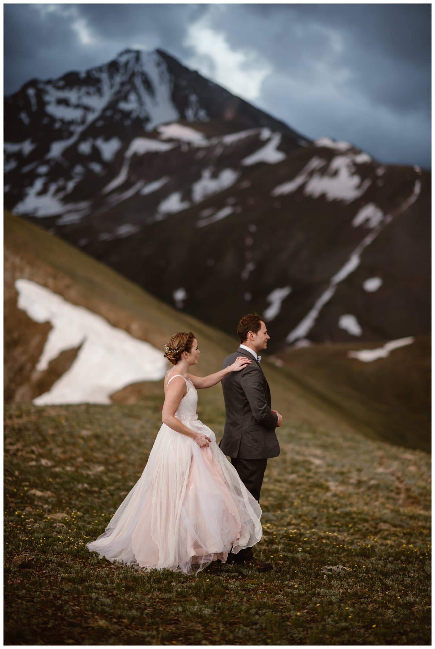Bride puts her hand on groom's shoulder as she stands behind him for a first look during a sunrise hiking elopement near Aspen, Colorado. 