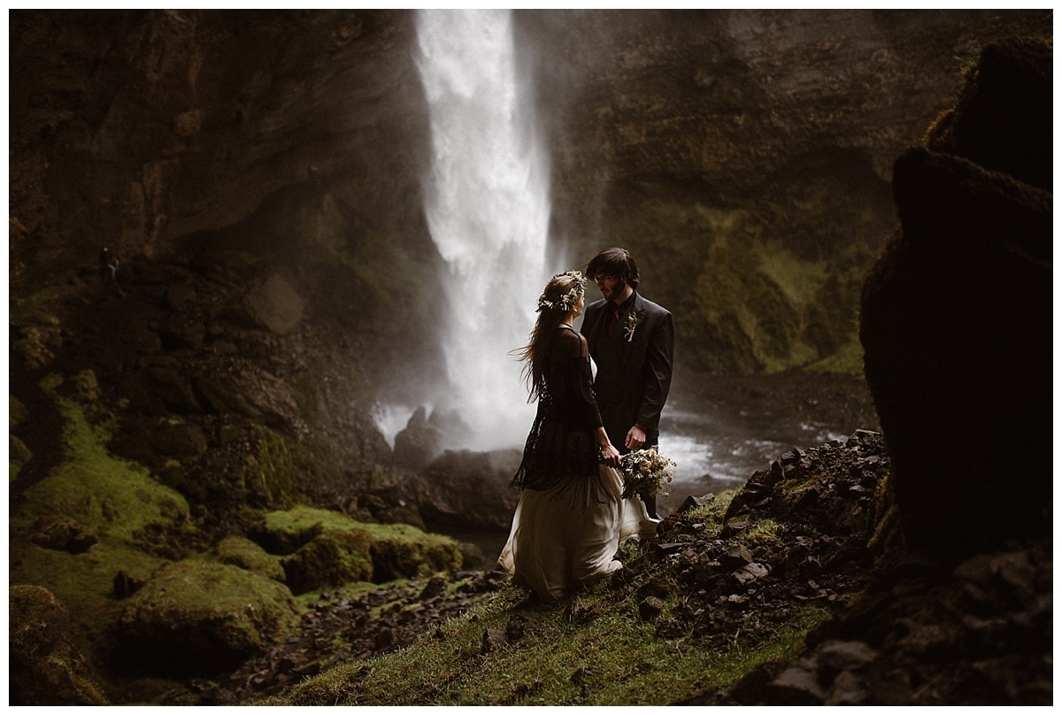 Bride and groom stand facing each other, in front of a waterfall, in Iceland. 