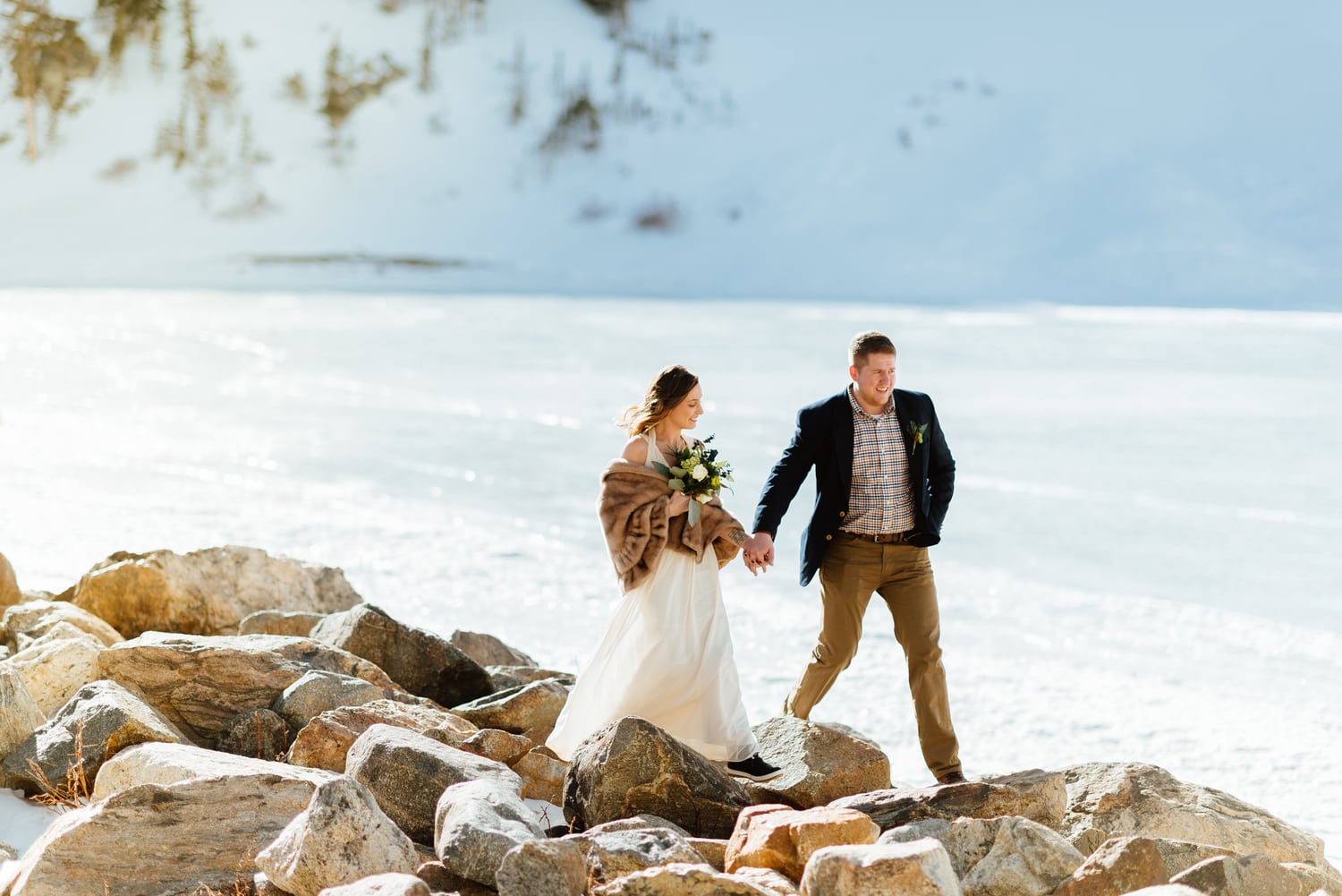 Bride and groom hold hands and walk across rocks, with snow covering the ground behind them at St. Mary's Glacier in Idaho Springs, Colorado. 