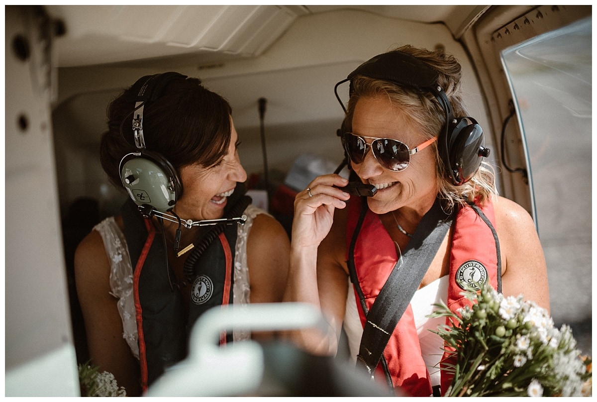 Two brides sitting in helicopter, smiling at each other on their elopement day in Tofino, Canada.