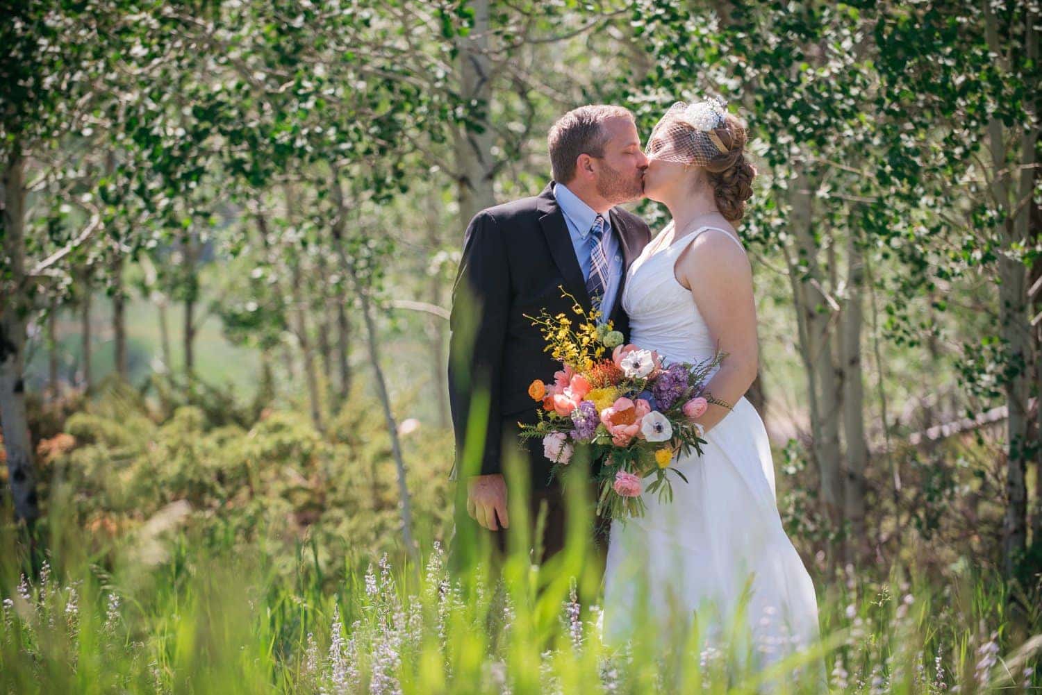 Bride and groom share a kiss on their elopement day in Grand Lake, Colorado. They are standing in a meadow with wildflowers and an aspen grove in the background. 