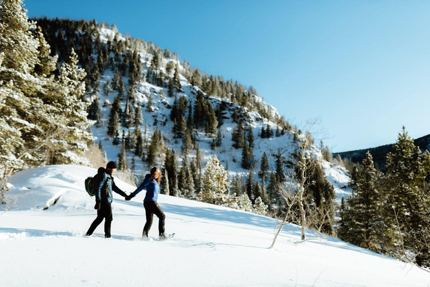 Couple holds hands while snowshoeing at Indian Peaks, in Colorado. There is snow covering the ground, and trees and mountains in the background. 