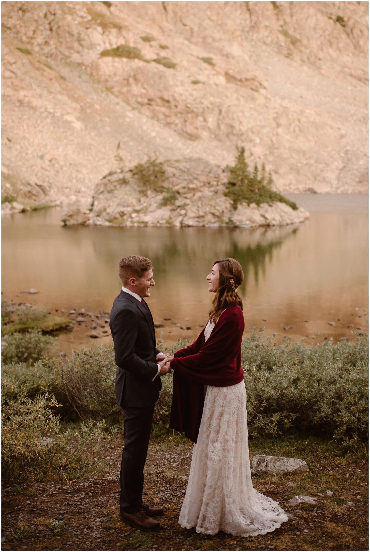 Bride and groom have first look in front of alpine lake with island in Buena Vista, Colorado. They are smiling at each other and holding hands. 