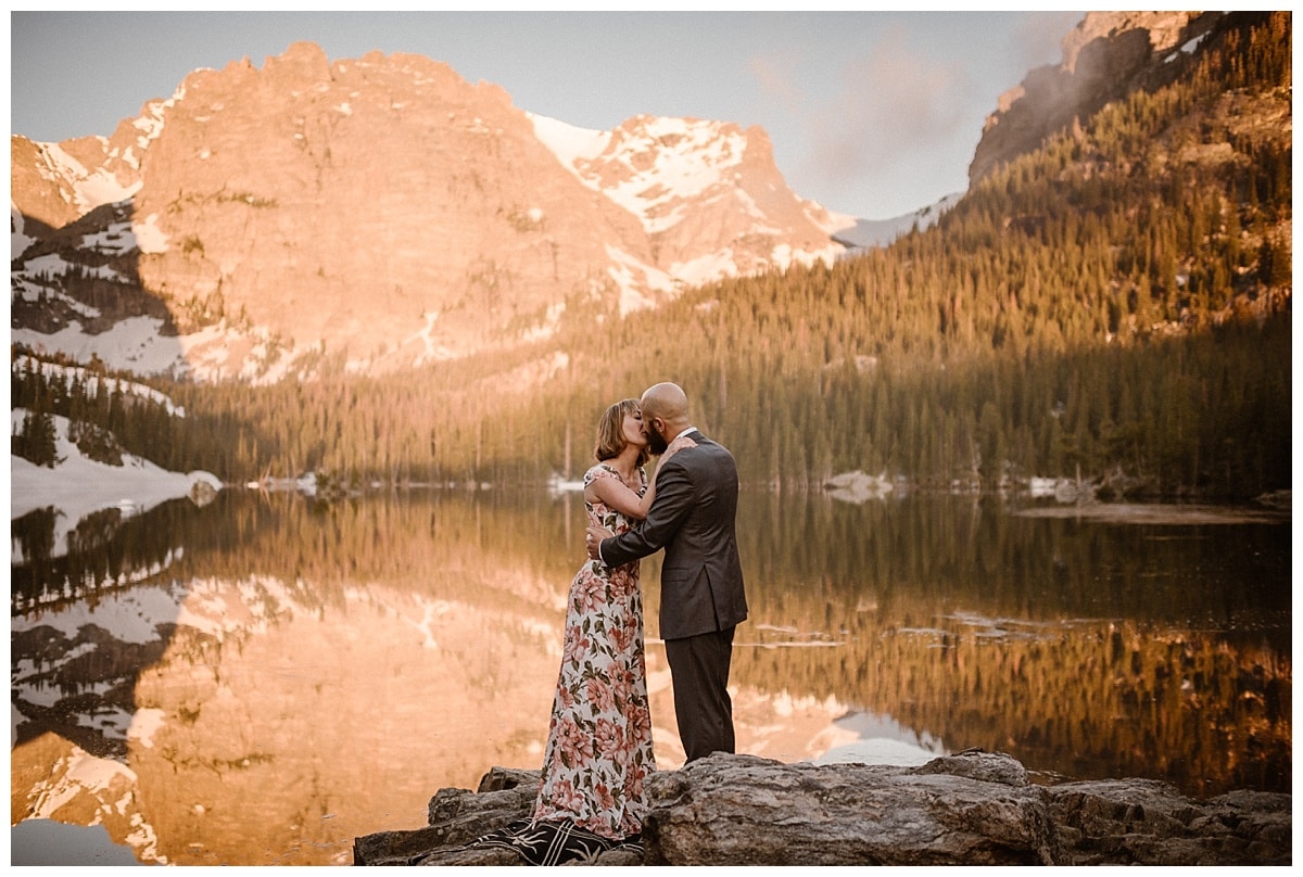 Bride and groom kiss during sunrise elopement in front of alpine lake. There is alpenglow on the mountains behind them. 