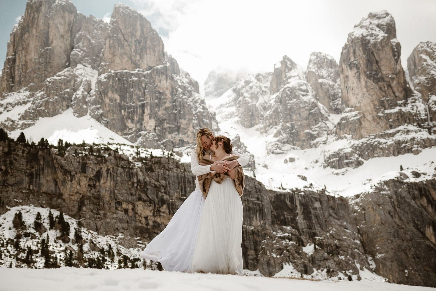 Two brides embrace in front of the snow-covered Italian Dolomites. 