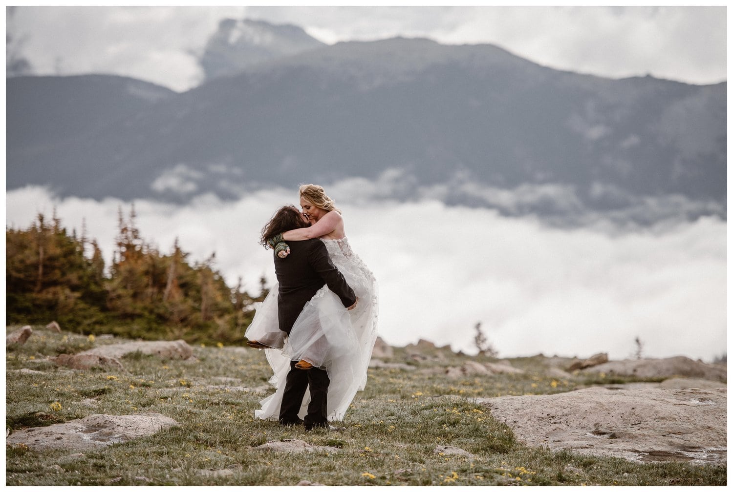 Groom lifts up bride as they share a kiss in a meadow at Trail Ridge Road in Rocky Mountain National Park, Colorado. There are cloud inversions behind them. 