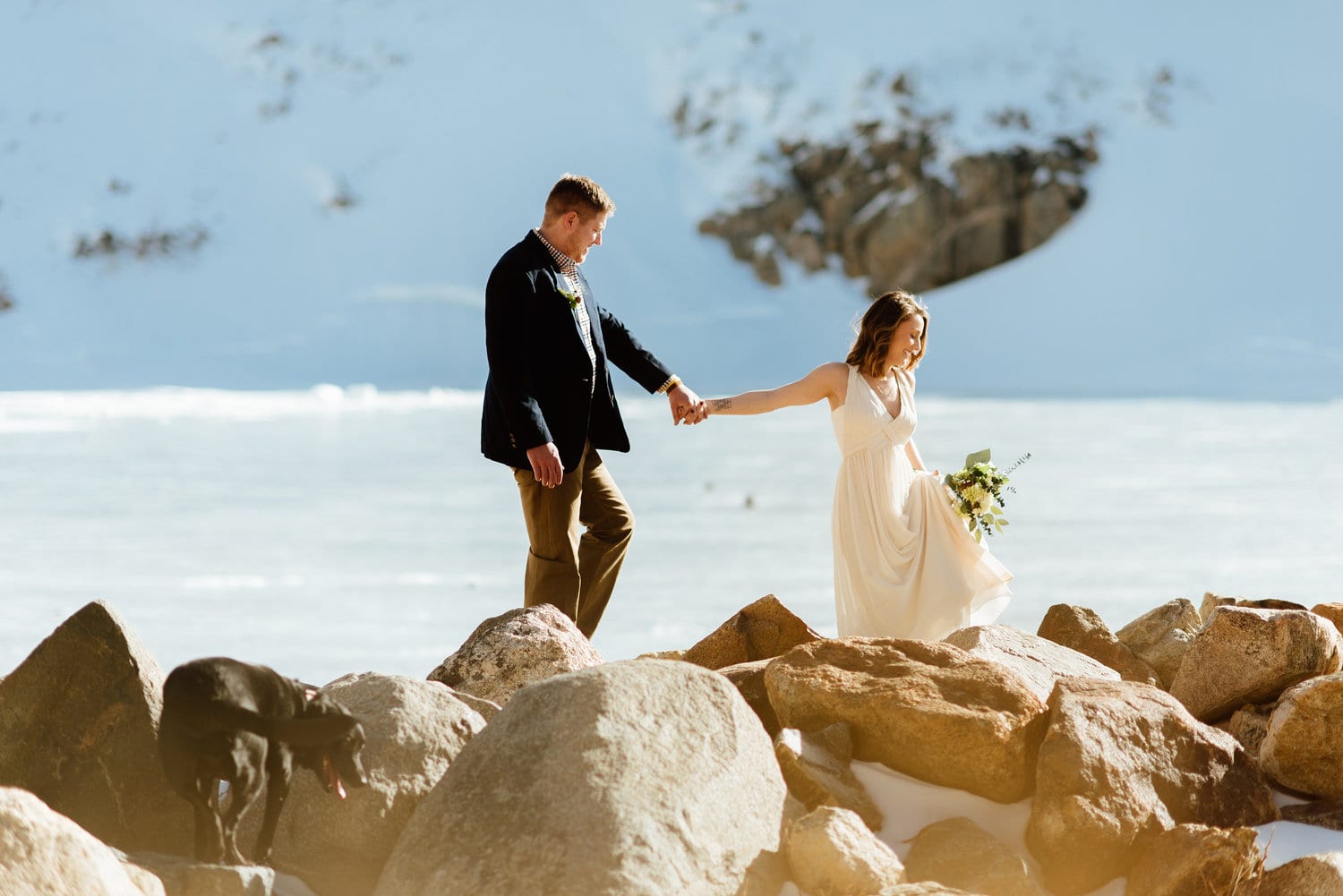 Bride and groom hold hands and walk across rocks, with their dog. Snow iscovering the ground behind them at St. Mary's Glacier in Idaho Springs, Colorado. 