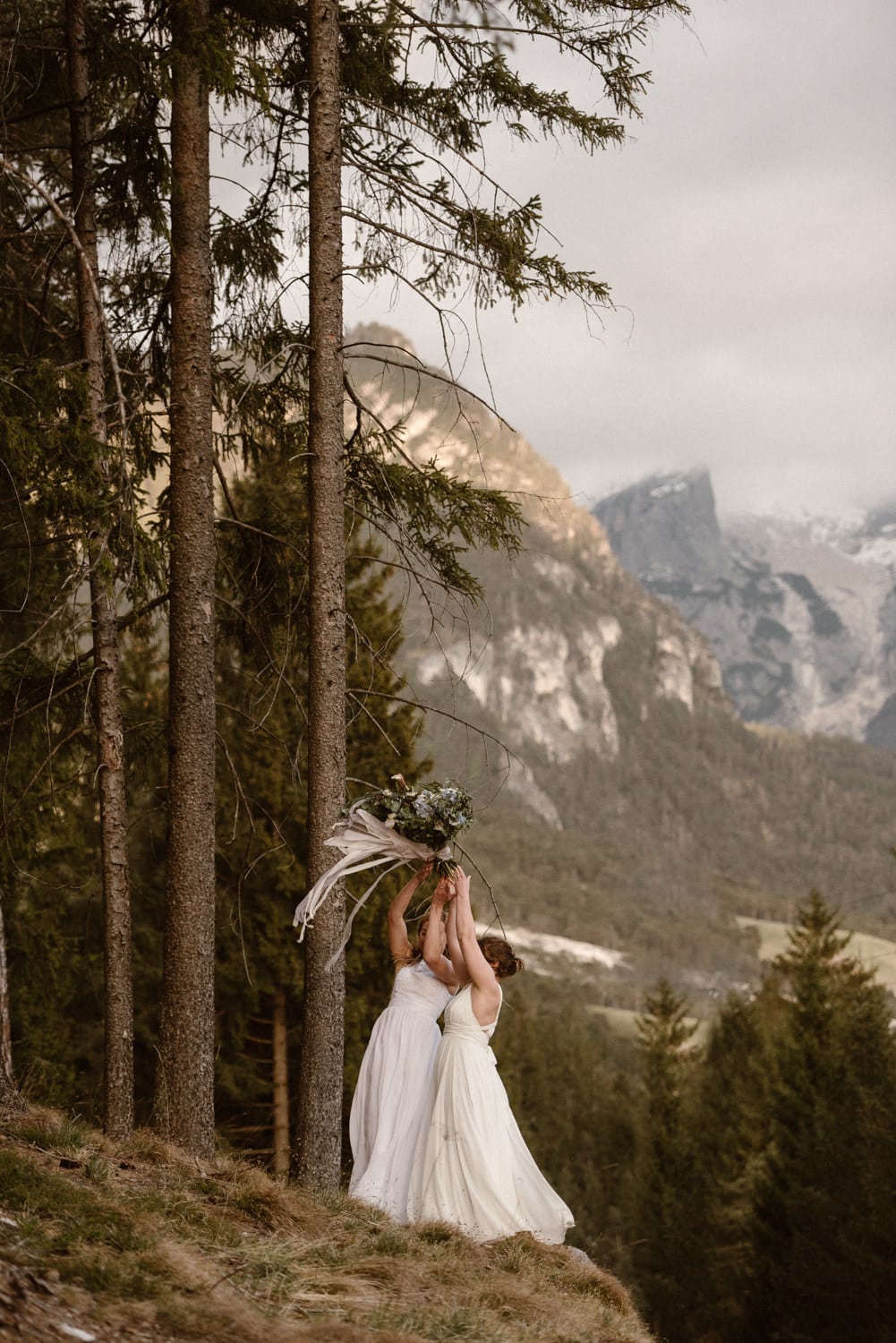 Two brides hold their bouquets in the air. They are both wearing white dresses and there are mountains in the background. 