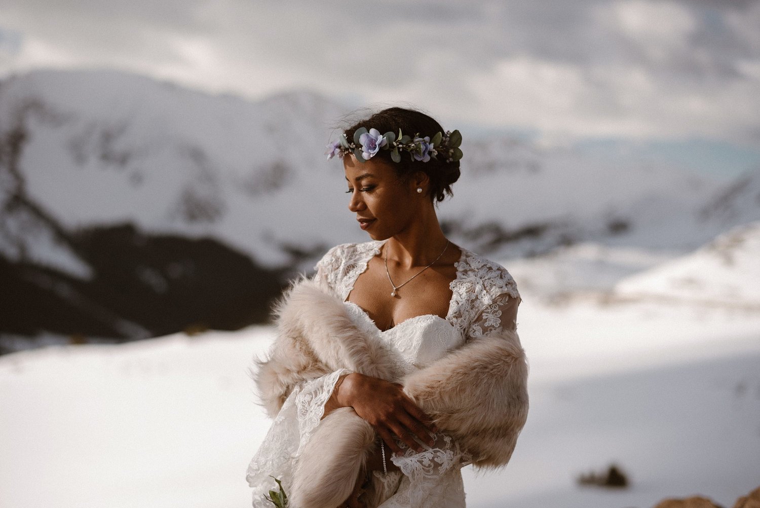 Bride on her elopement day at Loveland Pass, in Colorado. She is  looking down, wearing a purple flower crown, white dress, and a fur shawl. There are snow-capped mountains in the background. 
