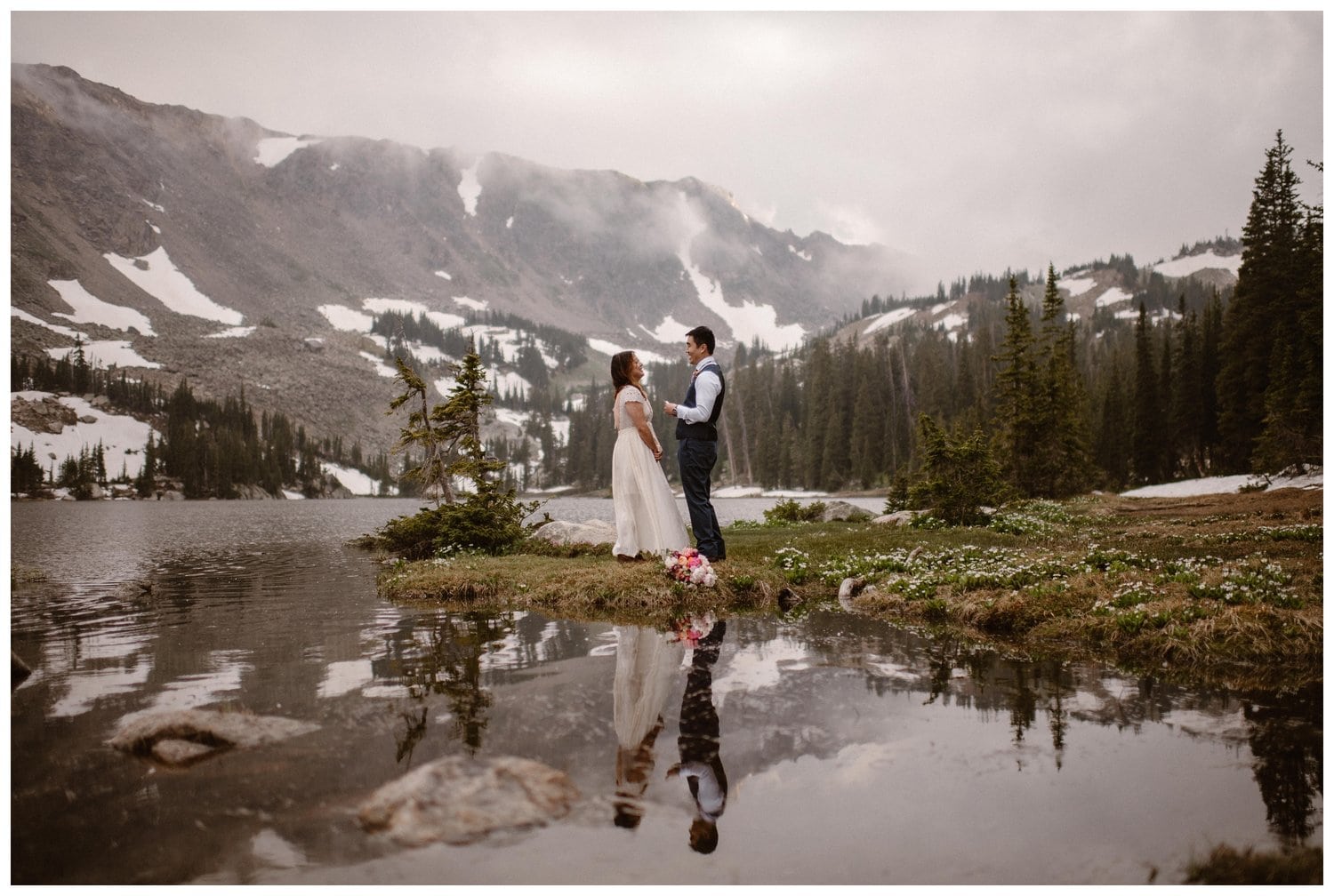 Bride and groom read their vows during intimate elopement ceremony in front of a high alpine lake, near Boulder, Colorado. There is a forest and snow-capped mountains in the background. 