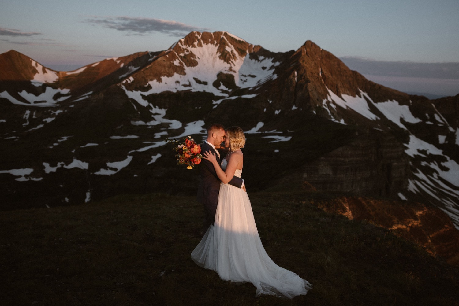 Bride and groom embrace and lean their foreheads against each other. There are snow-capped mountains behind them. 
