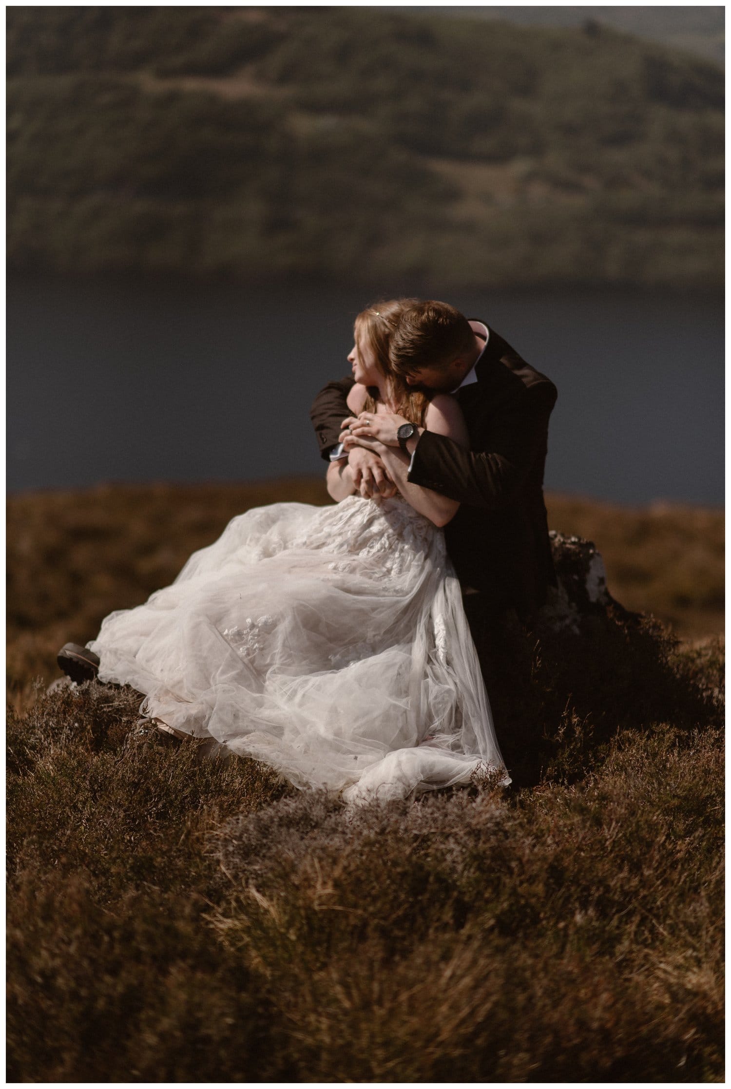 Groom has his arms wrapped around the bride, as they sit on a ridge overlooking Loch Ness, in the Scottish Highlands.
