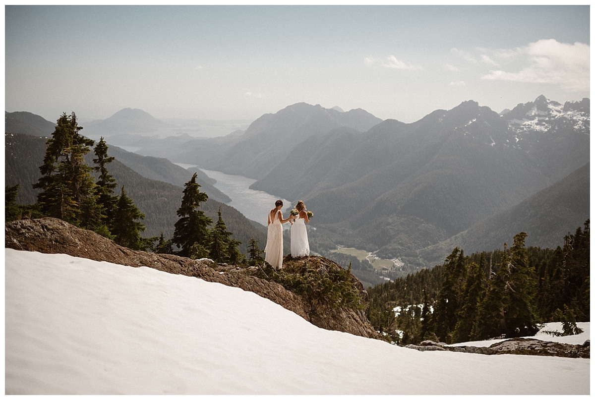 Two brides stand on cliff and look out at mountains in Tofino, Canada. There is snow on the ground around them. 
