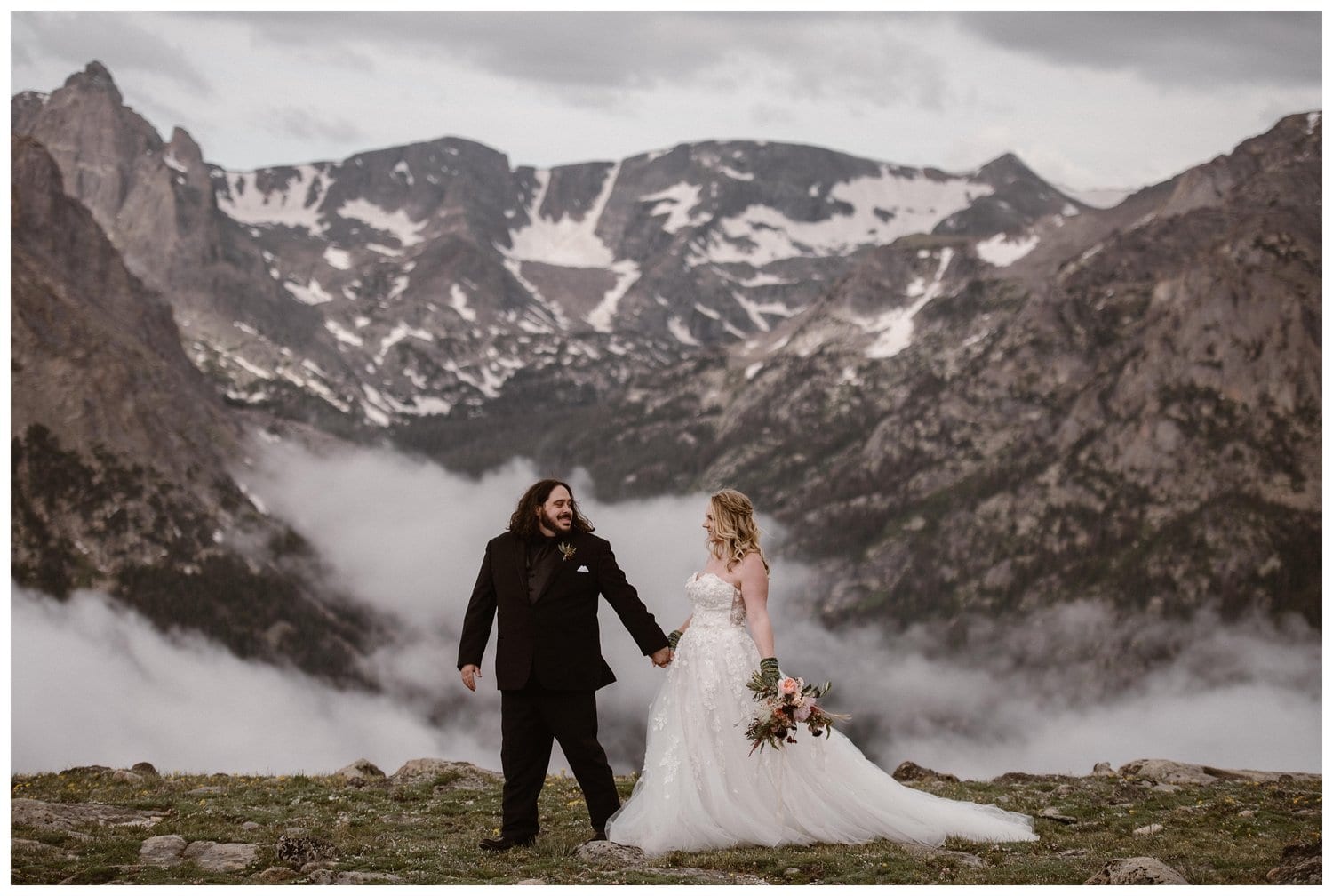Bride and groom hold hands and smile at each other. There are mountains and cloud inversions behind them at Trail Ridge Road in Rocky Mountain National Park, Colorado. 