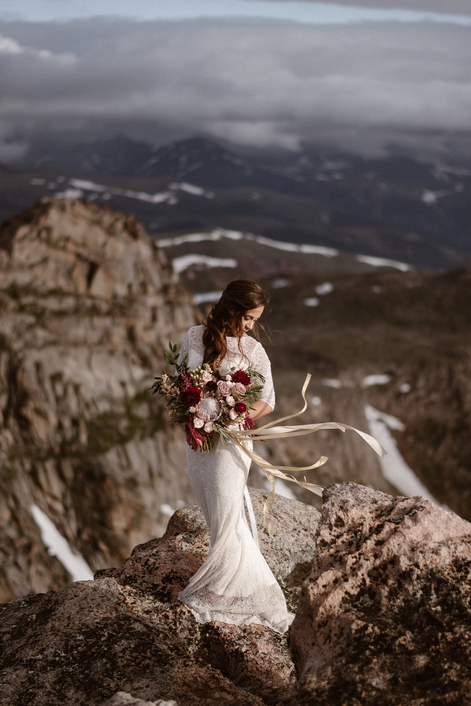Bride standing on a rocky cliff and looking down. She is wearing a white dress, and holding a bouquet with ribbons flowing in the wind. 
