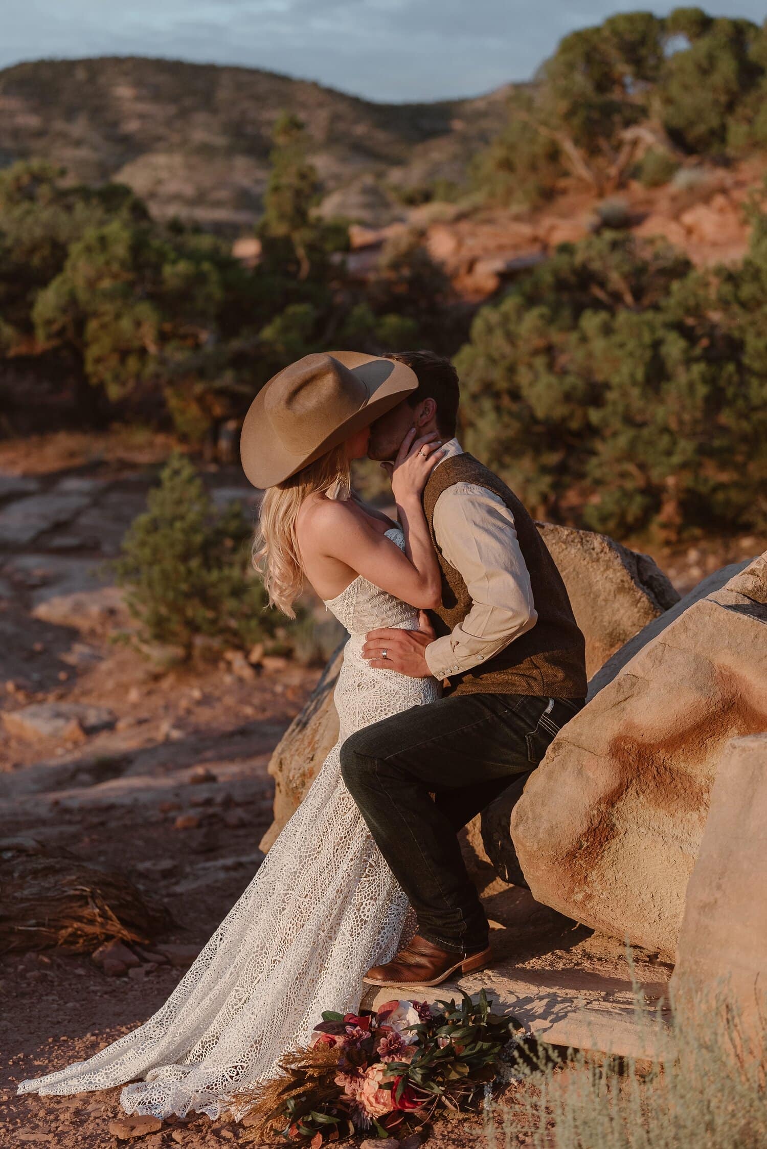 Bride and groom kiss while leaning against rock. Bride is wearing groom's cowboy hat. 