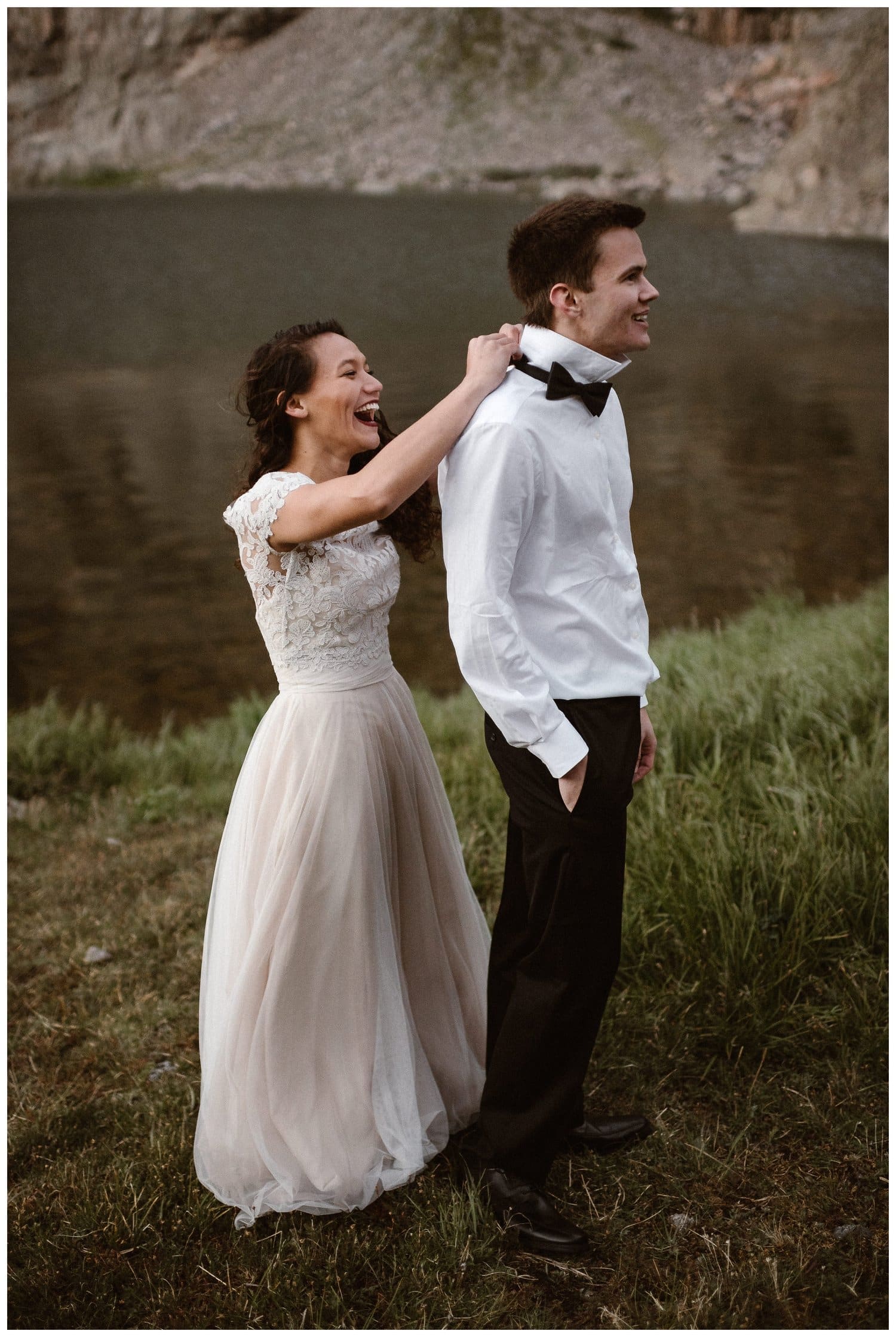 Bride helps groom put on his bowtie on their elopement day. They are both smiling, wearing wedding attire, and standing in front of a lake.