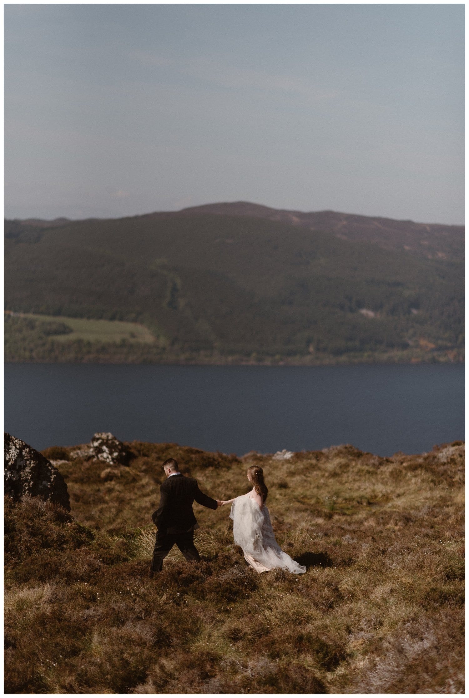 Bride and groom hold hands and hike around a ridge overlooking Loch Ness in the Scottish Highlands. 