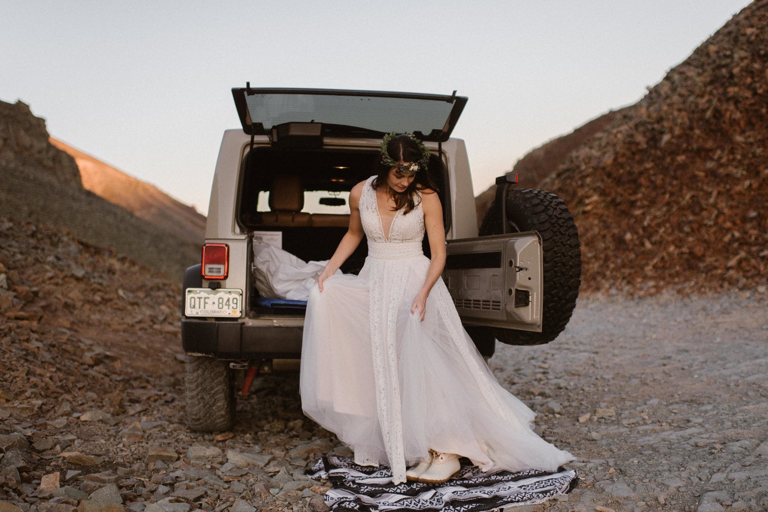 Bride getting ready for her elopement behind a Jeep. She is standing on a blanket and wearing a white dress and flower crown.