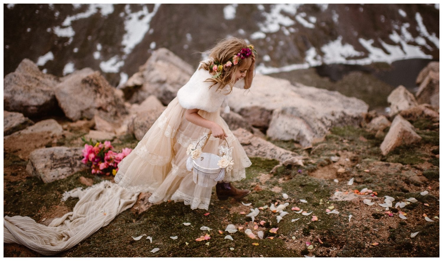 Young girl wearing a white dress, fur shawl, flower crown, and carrying a basket. There are flower petals creating a path in front of her, and snow-capped mountains in the background.