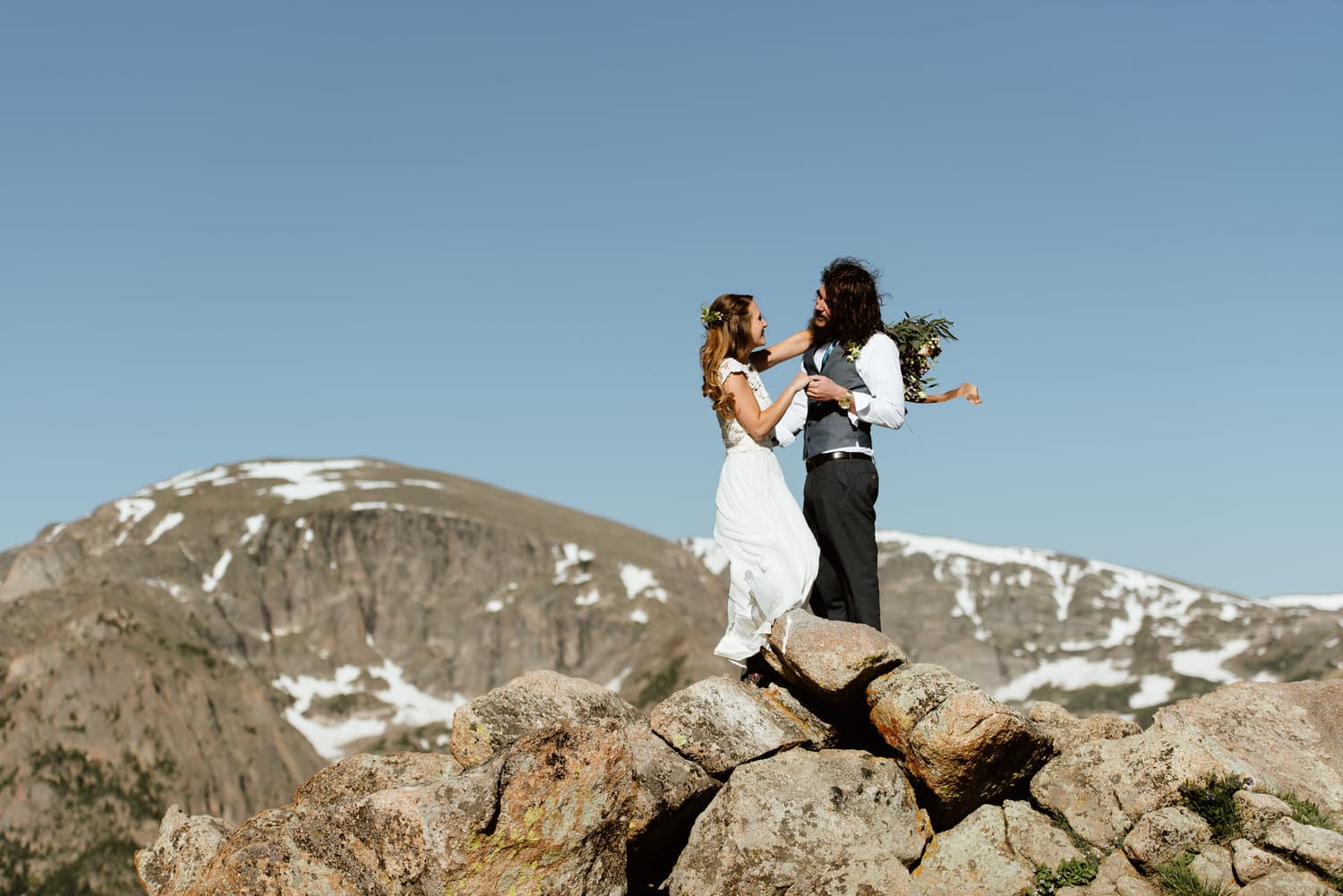 Bride and groom stand on rocks while holding hands and smiling at each other at Trail Ridge Road in Colorado. There are snow-capped mountains in the background. 