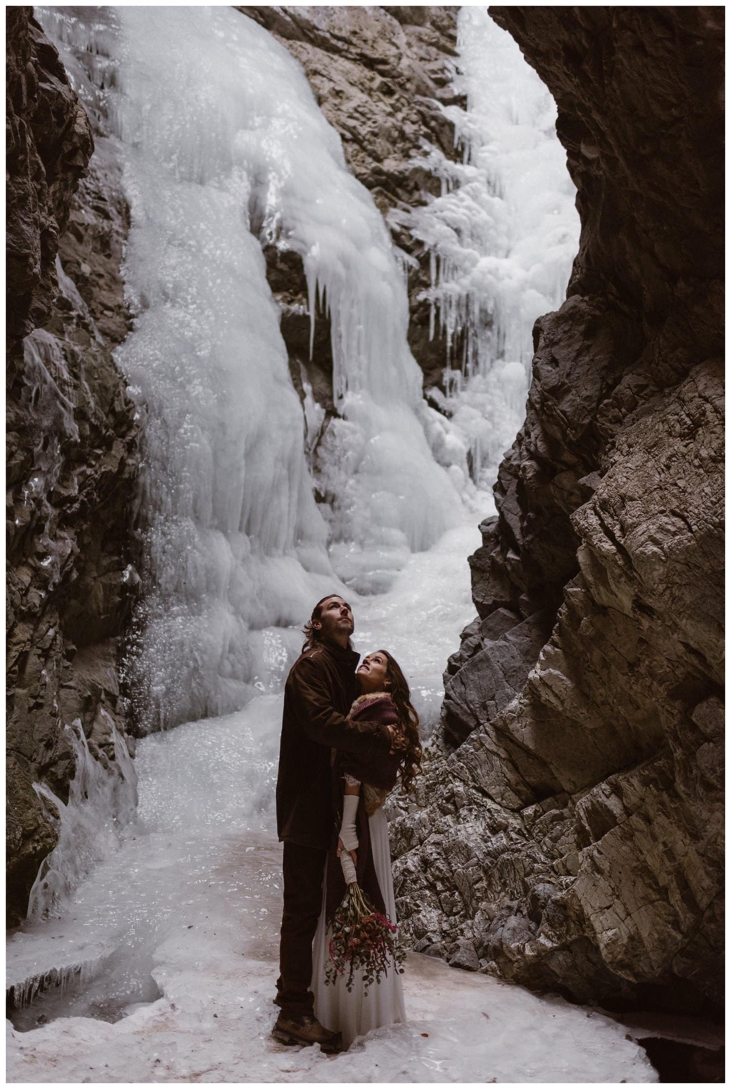 Bride and groom look up at frozen waterfall at Zapata Falls. 