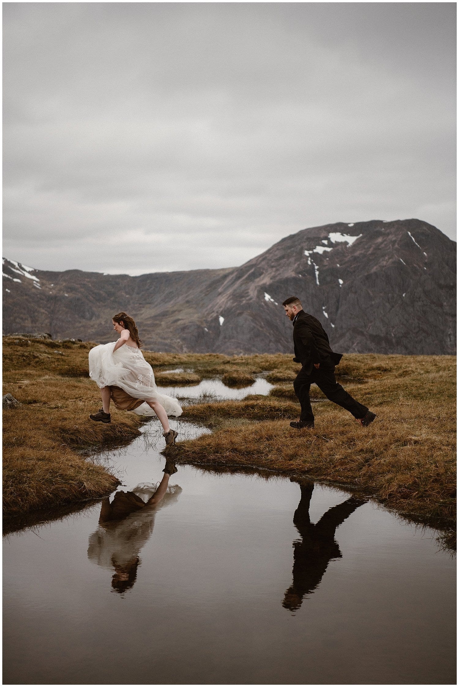 Bride and groom run, as bride leaps across puddle with mountains in background. 