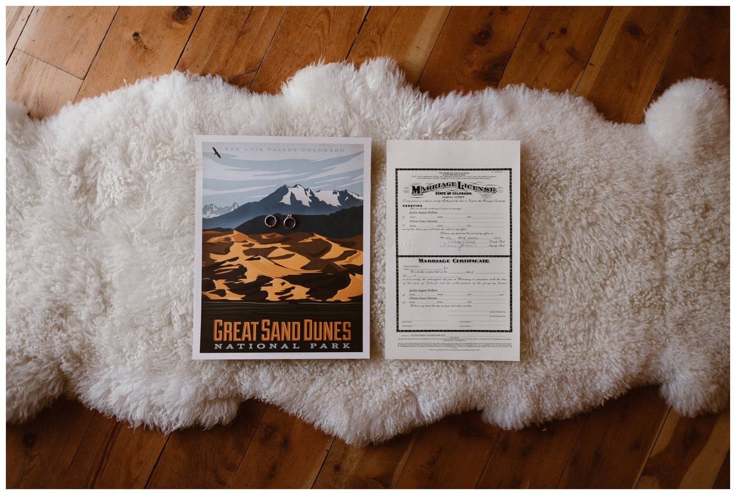 Flay lay of bride and groom's rings, marriage license, and poster of Great Sand Dunes National Park. 
