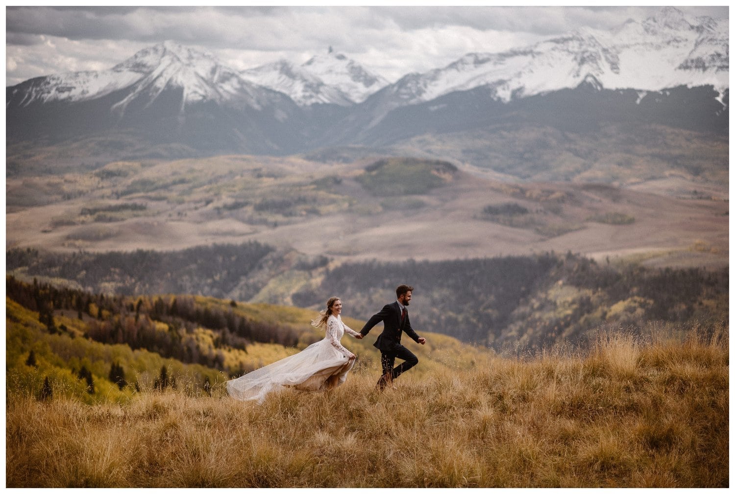 Bride and groom holding hands and running through a grassy meadow on their elopement day. There are snow-capped mountains in the background. 