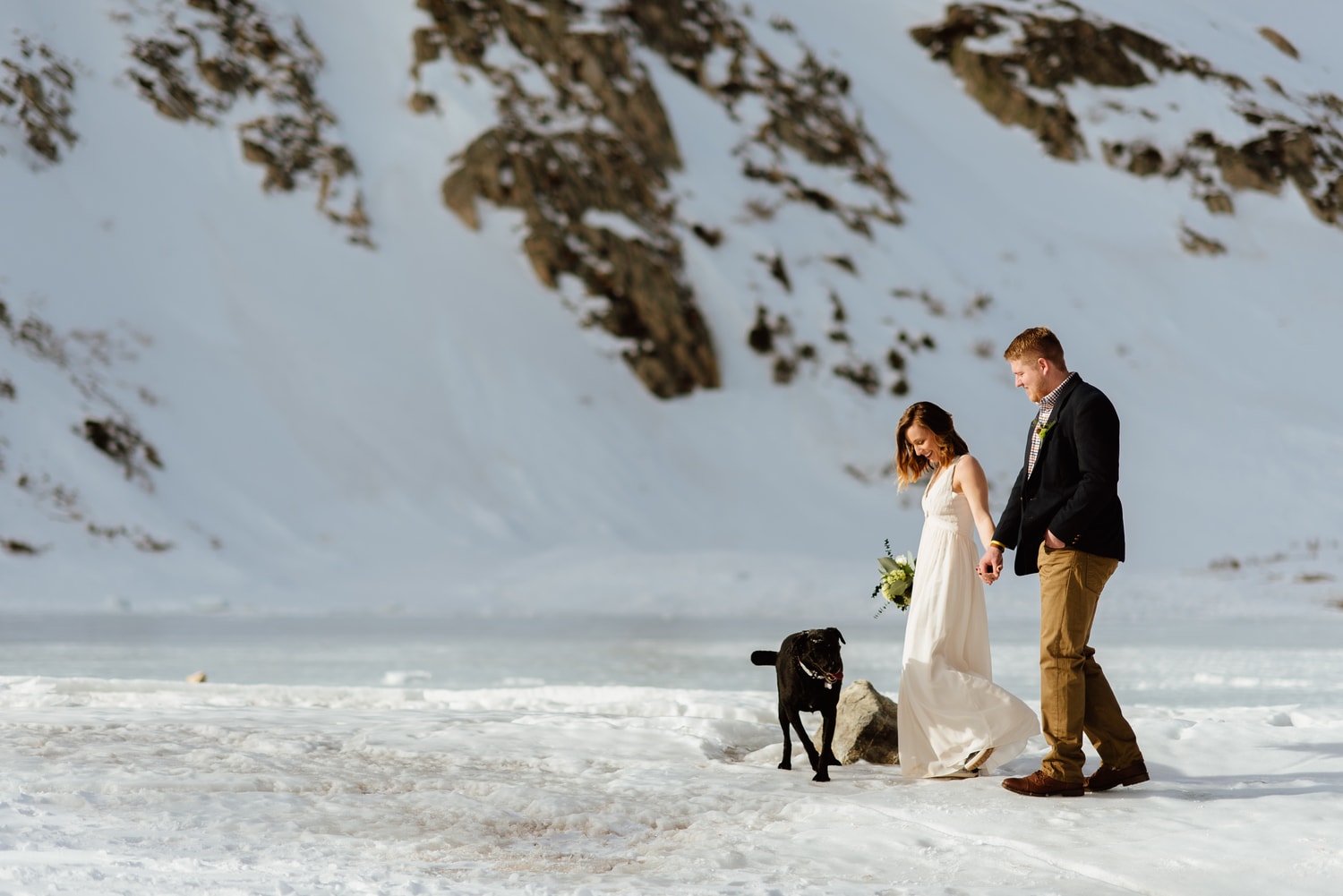 Bride and groom hold hands and walk through snow with their dog at St. Mary's Glacier in Idaho Springs, Colorado. 