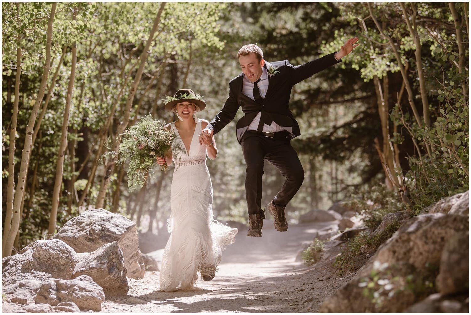 Bride and groom on hiking trail, surrounded by trees and rocks. Groom is jumping in the air. 