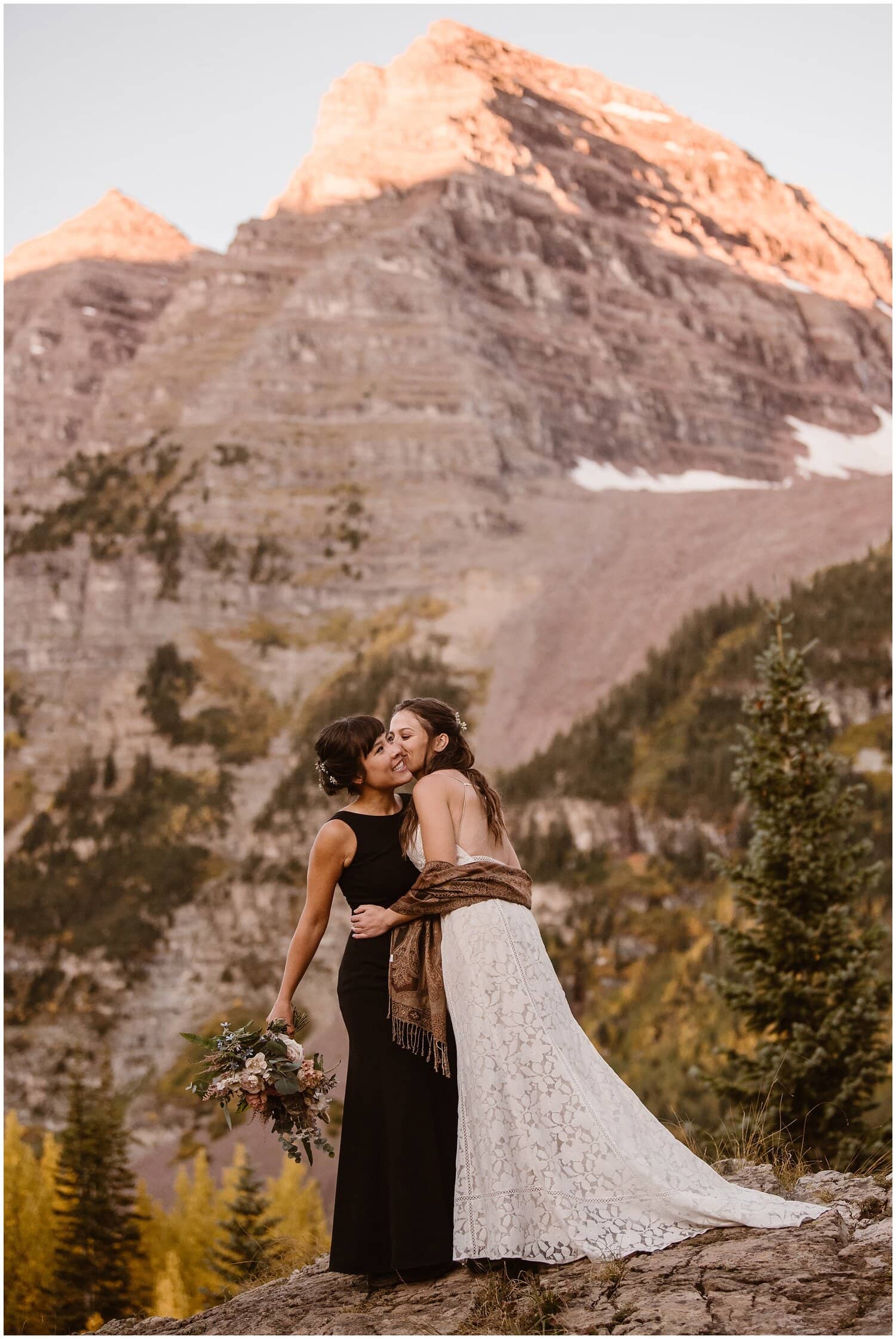 Two brides embrace. One kisses the other on the cheek. There are trees and mountains in the background. 