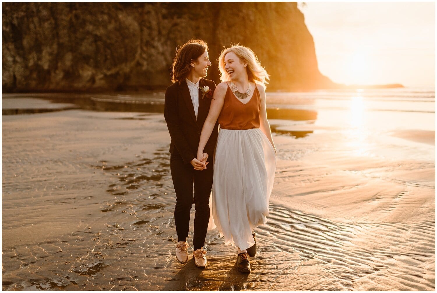 Two brides walk along the beach. They are holding hands and smiling at each other. 