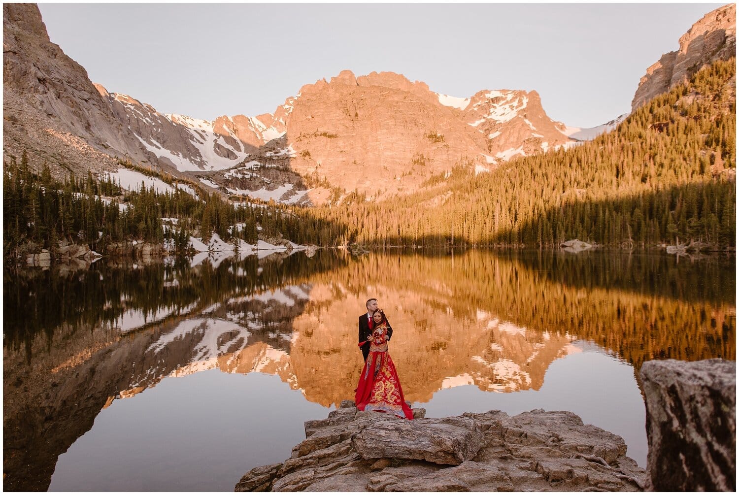 Bride and groom hug, while standing in front of an alpine lake. Alpenglow on the mountains behind them. 